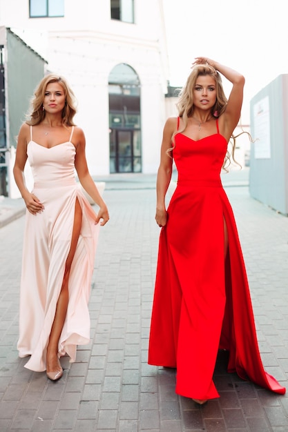 Duas garotas elegantes e lindas usando vestidos de noite elegantes Mulheres atraentes esbeltas com cabelos longos loiros andando e olhando para a câmera Garotas posando em vestidos longos vermelhos e em pó