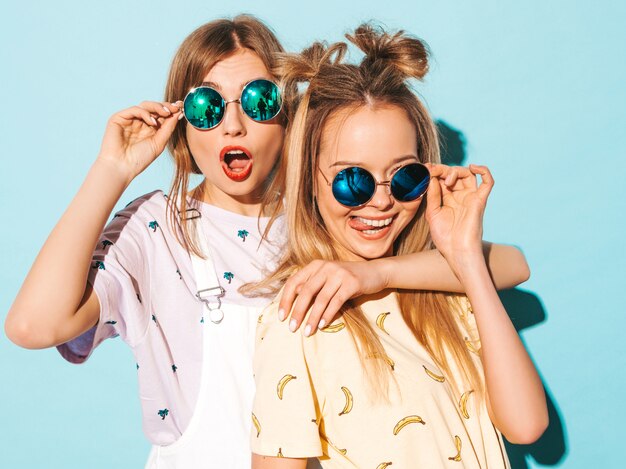 Duas garotas loiras hipster sorrindo lindas jovens em roupas de camiseta colorida na moda verão. e mostrando a língua