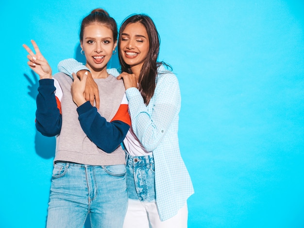 Duas garotas loiras hipster sorrindo lindas jovens em roupas de camiseta colorida na moda verão. Mulheres despreocupadas "sexy" que levantam perto da parede azul. Modelos positivos se divertindo