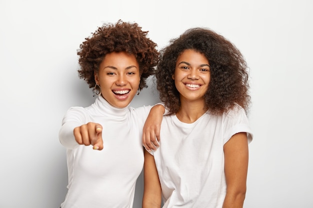 Duas garotas milenares amigáveis e felizes têm rostos felizes, ficam lado a lado, apontam para a distância