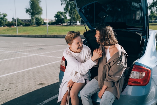 Duas garotas no estacionamento no porta-malas aberto posando para a câmera.