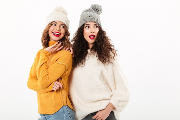 Duas garotas sorridentes em blusas e chapéus juntos enquanto olha para longe sobre parede branca