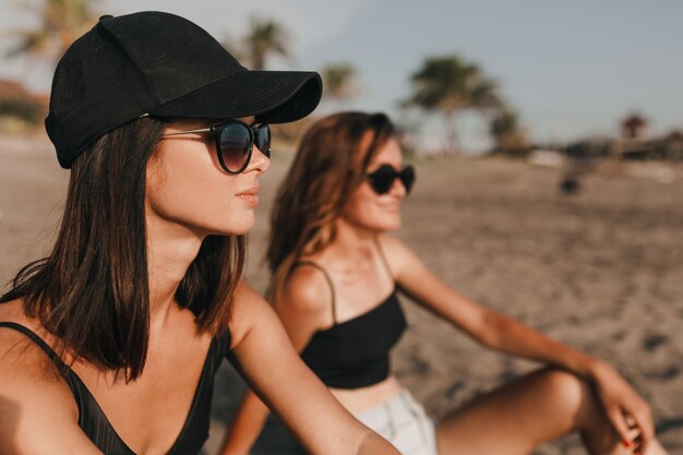 Duas garotas usando óculos escuros e maiôs sentadas na praia com palmeiras e olhando para o mar com sorrisos românticos Duas melhores amigas passam as férias nos trópicos