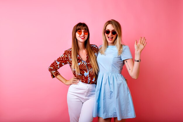 Duas irmãs bonitas felizes, melhores amigas, mulheres hipster se divertindo juntas no fundo rosa do estúdio