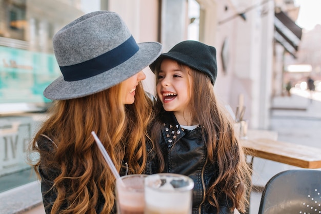 Duas irmãs encaracoladas de cabelos compridos, olhando com amor um para o outro, aproveitando a manhã ensolarada no café ao ar livre.