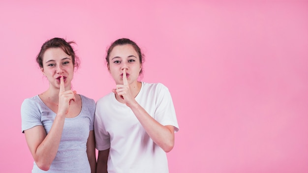 Duas irmãs fazendo gesto quieto contra pano de fundo rosa