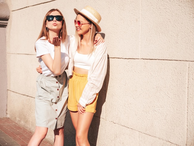 Duas jovem bela mulher hipster sorridente em roupas da moda de verão Mulheres despreocupadas sexy posando na rua com chapéu Modelos puros positivos se divertindo ao pôr do sol