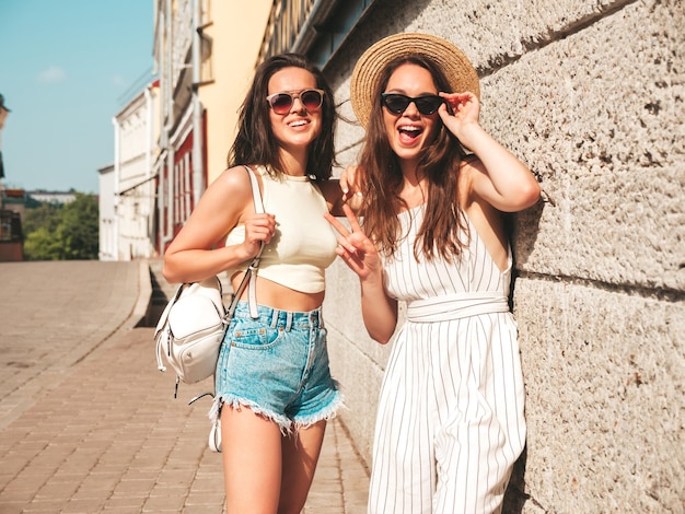 Duas jovem bela mulher hipster sorridente em roupas da moda de verãomulheres despreocupadas sexy posando no fundo da rua no chapéu modelos puros positivos se divertindo ao pôr do sol abraçando