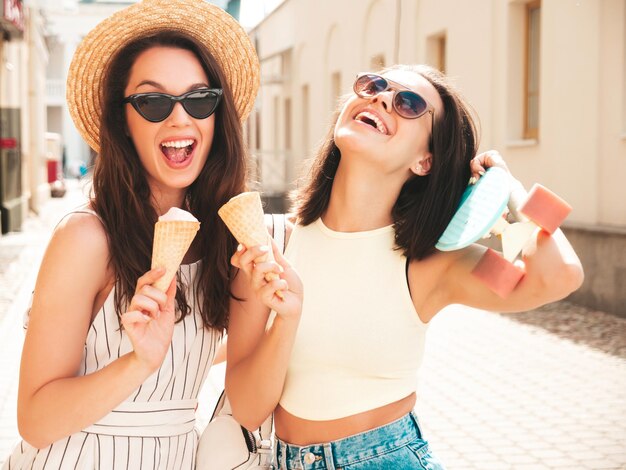 Duas jovem e bela mulher de hipster sorridente em roupas da moda de verão e vestidoMulheres despreocupadas sexy posando no fundo da rua Modelos positivos comendo sorvete saboroso no cone de waffles ao pôr do sol