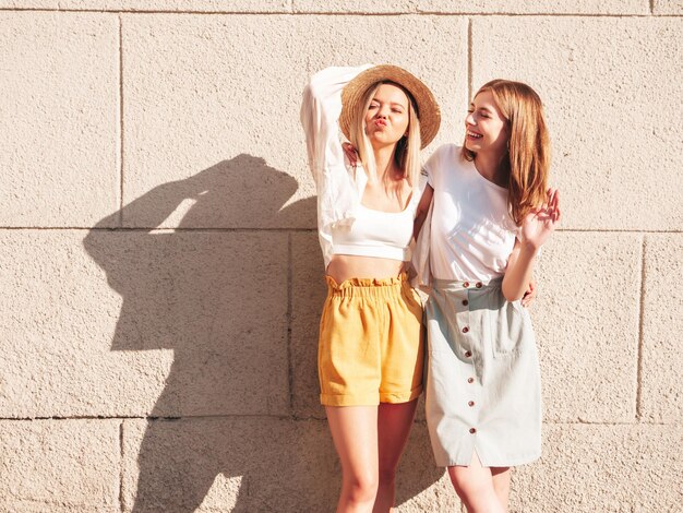 Duas jovem e bela mulher hipster sorridente em roupas da moda de verãoMulheres despreocupadas sexy posando perto de parede branca na rua Modelos puros positivos se divertindo ao pôr do sol abraçando e enlouquecendo