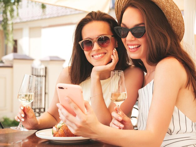 Duas jovem e bela mulher hipster sorridente em roupas de verãoMulheres despreocupadas posando no café da varanda na ruaModelos positivos bebendo videira branca no chapéuOlhando para a tela do smartphone usando aplicativos