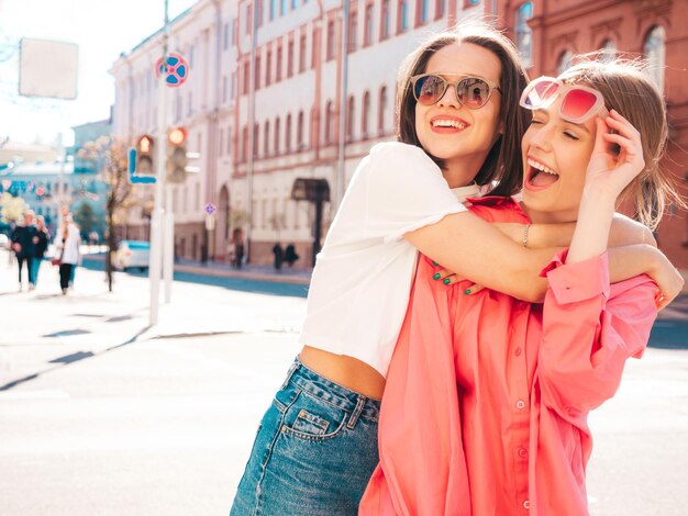 Duas jovem linda sorridente hipster feminina em jeans e roupas de t-shirt branca na moda verão. Mulheres despreocupadas sexy posando no fundo da rua. Modelos positivos se divertindo, abraçando e enlouquecendo