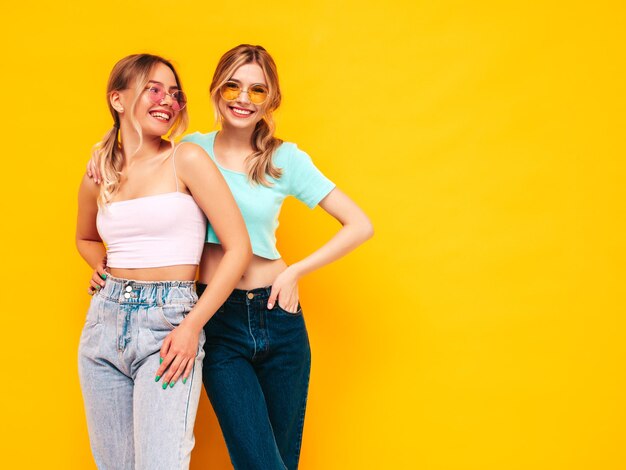 Duas jovem linda sorridente loira hipster em roupas da moda de verão Mulheres despreocupadas sensuais posando perto da parede amarela no estúdio Modelos positivos se divertindo Alegre e feliz em óculos de sol