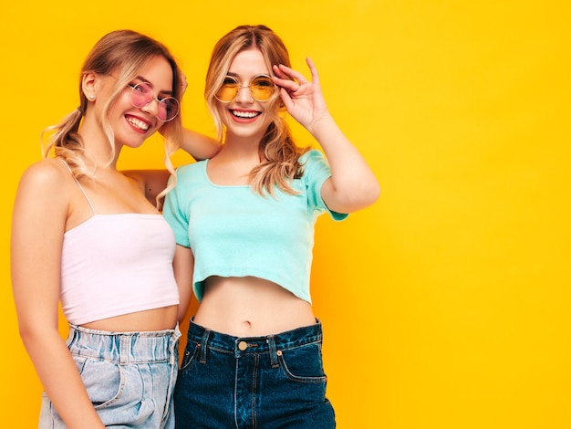 Duas jovem linda sorridente loira hipster em roupas da moda de verão Mulheres despreocupadas sensuais posando perto da parede amarela no estúdio Modelos positivos se divertindo Alegre e feliz em óculos de sol