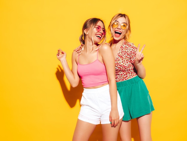 Duas jovem linda sorridente loira hipster em roupas da moda de verão mulheres despreocupadas sensuais posando perto da parede amarela no estúdio modelos positivos se divertindo alegre e feliz em óculos de sol