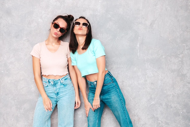 Foto grátis duas jovem linda sorridente loira hipster em roupas da moda de verão mulheres despreocupadas sensuais posando perto da parede cinza no estúdio modelos positivos se divertindo alegre e feliz em óculos de sol