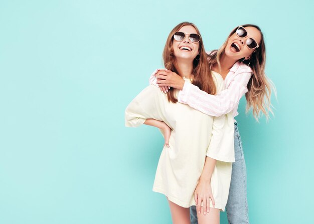 Duas jovem linda sorridente morena hipster feminina em roupas da moda de verão Mulheres despreocupadas sensuais posando perto da parede azul Modelos positivos se divertindo Alegre e feliz em óculos de sol