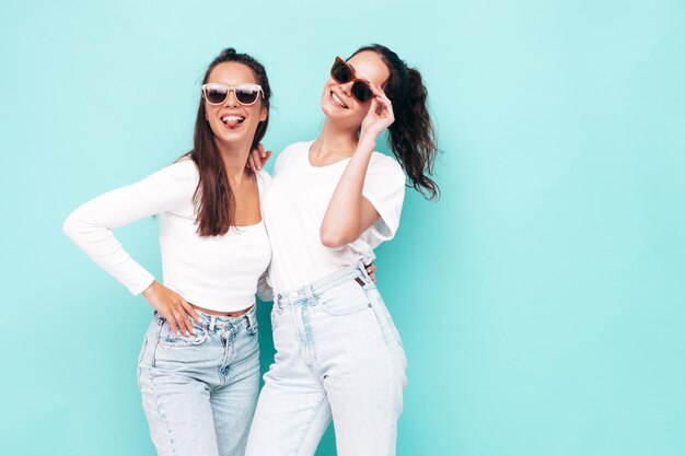 Duas jovem linda sorridente morena hipster feminina em roupas da moda de verão Mulheres despreocupadas sensuais posando perto da parede azul Modelos positivos se divertindo Alegre e feliz em óculos de sol