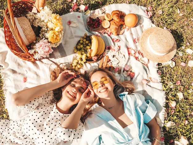 Duas jovens belas mulheres sorridentes no vestido de verão da moda e chapéus. Mulheres despreocupadas fazendo piquenique do lado de fora.