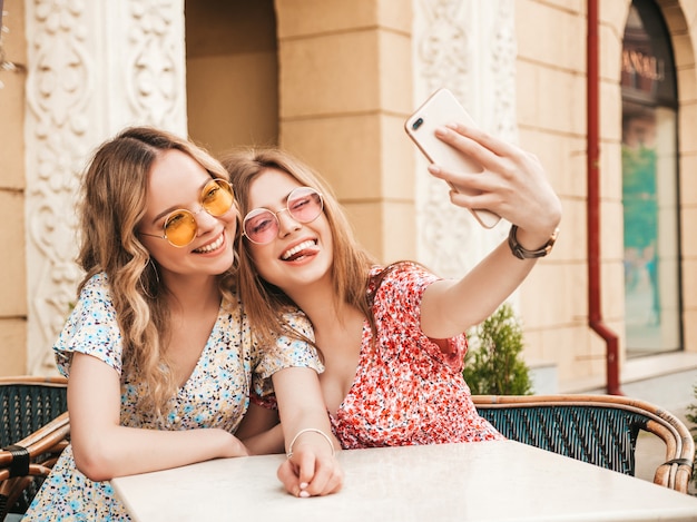 Duas jovens bonitas garotas hipster sorridentes no vestido de verão na moda. Mulheres despreocupadas conversando no café varanda na rua fundo. Modelos positivos se divertindo e tomando selfie no smartphone