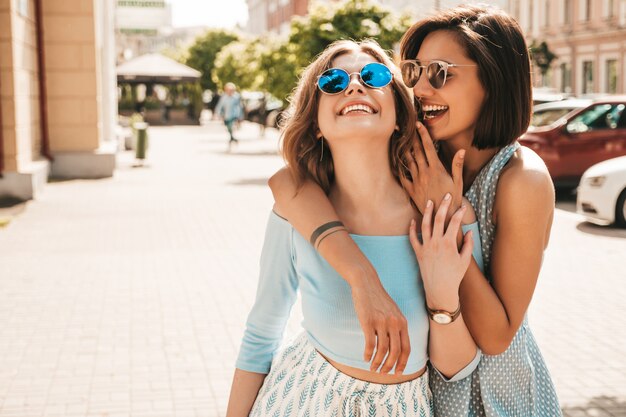 Duas jovens bonitas hipster garotas sorridentes em roupas da moda no verão. Mulheres despreocupadas sexy posando na rua fundo em óculos de sol. Modelos positivos se divertindo e abraçando