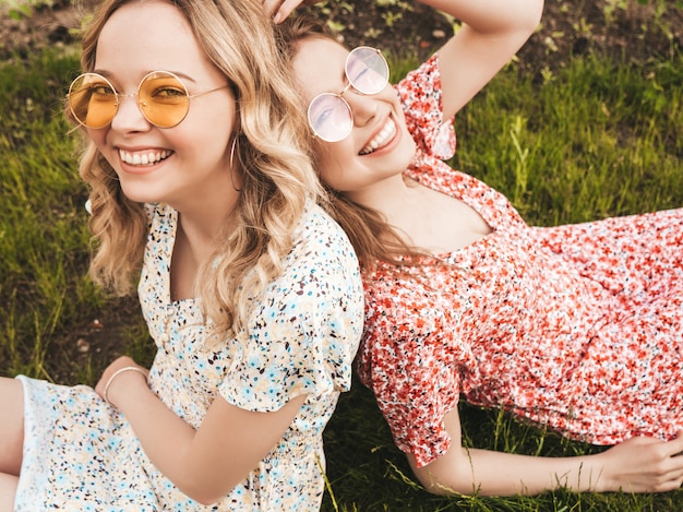 Duas jovens bonitas hipster garotas sorridentes no vestido de verão na moda. Mulheres despreocupadas sexy deitado na grama verde em óculos de sol. Modelos positivos se divertindo. Vista superior