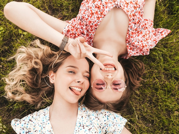 Duas jovens bonitas hipster garotas sorridentes no vestido de verão na moda. Mulheres despreocupadas sexy deitado na grama verde em óculos de sol. Modelos positivos se divertindo. Vista superior