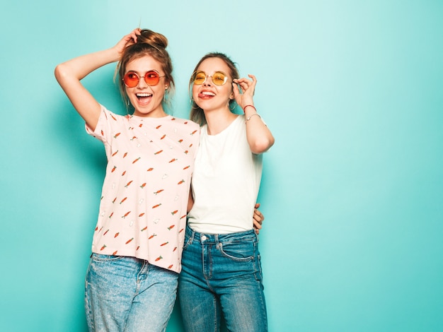 Duas jovens bonitas loiras sorridentes garotas hipster em roupas da moda verão hipster jeans. Mulheres despreocupadas "sexy" que levantam perto da parede azul. Modelos modernos e positivos se divertindo em óculos de sol
