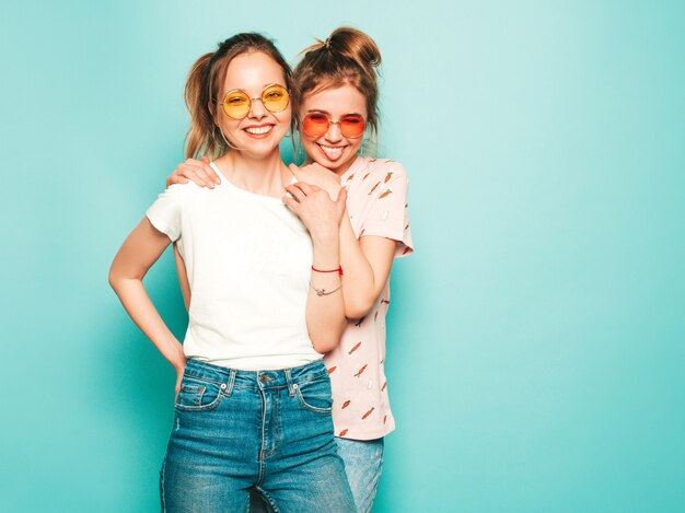 Duas jovens bonitas loiras sorridentes garotas hipster em roupas da moda verão hipster jeans. Mulheres despreocupadas "sexy" que levantam perto da parede azul. Modelos modernos e positivos se divertindo