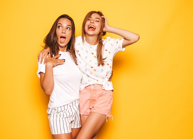 Duas jovens bonitas sorridentes garotas hipster em roupas da moda no verão. Mulheres despreocupadas "sexy" que levantam perto da parede amarela. Modelos positivos enlouquecendo e se divertindo.