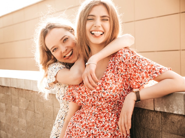 Duas jovens bonitas sorrindo garotas hipster em vestido de verão na moda. Mulheres despreocupadas sexy posando no fundo da rua. Modelos positivos se divertindo e mostrando línguas