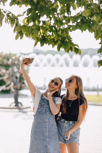 Duas jovens garotas bonitas em uma caminhada no parque tirando fotos de si mesmos no telefone. em um dia ensolarado de verão, alegria e amizades.