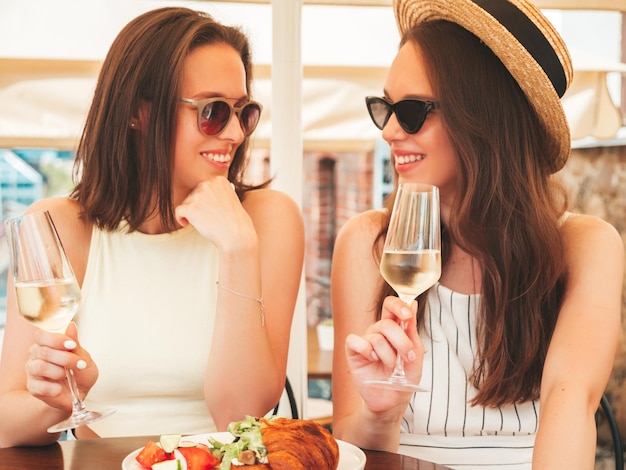 Duas jovens lindas e sorridentes hipster femininas em roupas da moda de verãoMulheres despreocupadas posando no café da varanda na ruaModelos positivos bebendo videira branca no chapéu Aproveitando suas férias