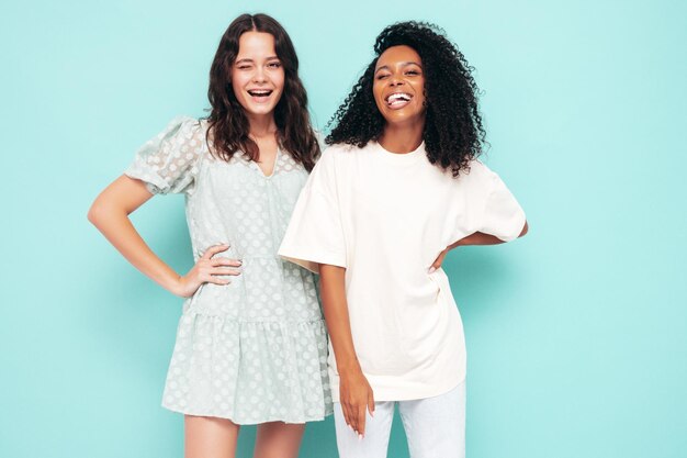 Duas jovens lindas e sorridentes hipster internacional em roupas da moda de verão Mulheres despreocupadas sensuais posando perto da parede azul no estúdio Modelos positivos se divertindo Conceito de amizade