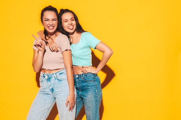 Duas jovens lindas e sorridentes hipster loira em roupas da moda de verão Mulheres despreocupadas sensuais posando perto da parede amarela no estúdio Modelos positivos se divertindo Alegre e feliz