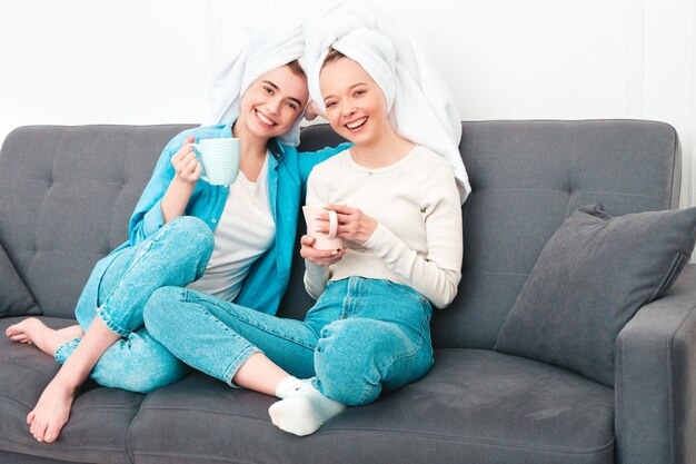 Duas jovens lindas mulheres sorridentes, sentadas no sofá. modelos despreocupados posando dentro de casa em um apartamento chique ou quarto de hotel. Eles fazem tratamentos de beleza em casa