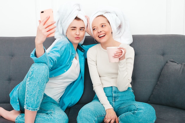 Duas jovens lindas mulheres sorridentes, sentadas no sofá. modelos despreocupados posando em ambientes fechados em um apartamento chique ou quarto de hotel