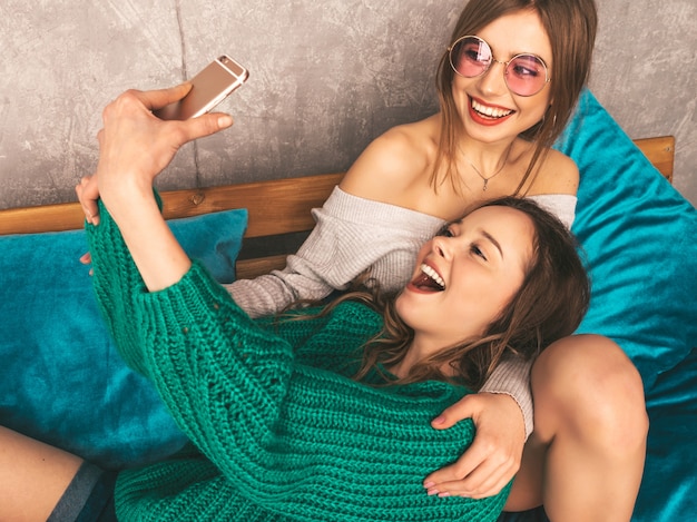 Duas jovens lindas sorrindo lindas garotas em roupas da moda no verão. Mulheres despreocupadas sexy, posando no interior e tomar selfie. Modelos positivos se divertindo com o smartphone.