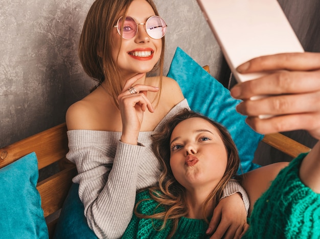 Duas jovens lindas sorrindo lindas garotas em roupas da moda no verão. mulheres despreocupadas sexy, posando no interior e tomar selfie. modelos positivos se divertindo com o smartphone.