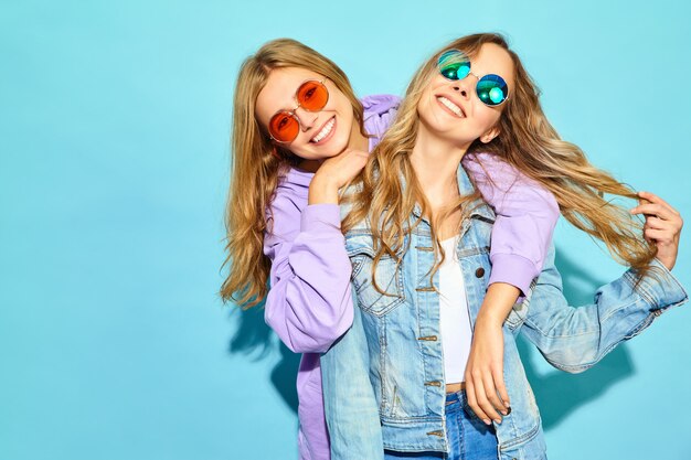 Duas jovens loiras lindas hipster mulheres sorridentes em roupas da moda no verão. Mulheres despreocupadas "sexy" que levantam perto da parede azul nos óculos de sol. Modelos positivos enlouquecendo e abraçando