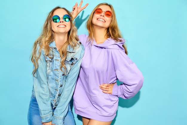 Duas jovens loiras lindas hipster mulheres sorridentes em roupas da moda no verão. Mulheres despreocupadas "sexy" que levantam perto da parede azul nos óculos de sol. Modelos positivos enlouquecendo