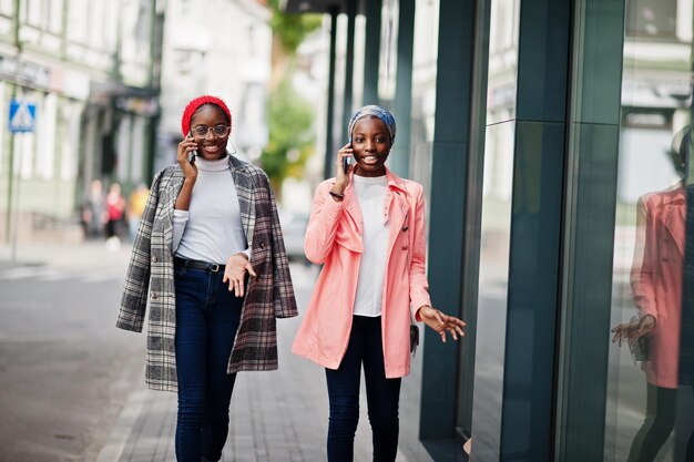 Duas jovens mulheres muçulmanas africanas, altas e magras, atraentes, modernas, em hijab ou lenço de cabeça de turbante e casaco posaram com telefones celulares