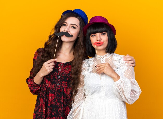 Duas jovens mulheres satisfeitas com um chapéu de festa, olhando para a frente, segurando bigode e lábios falsos