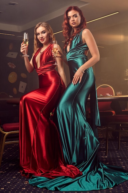 Duas jovens senhoras sensuais em longos vestidos de cetim, com champanhe nas mãos estão posando de lado contra uma mesa de pôquer no cassino de luxo. Paixão, cartas, fichas, álcool, vitória, jogo - é uma mulher