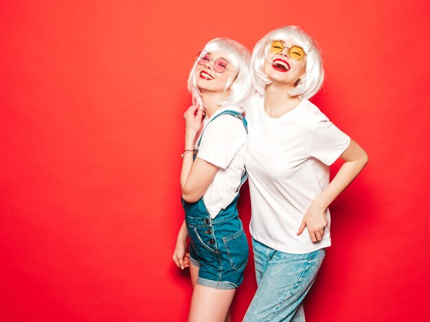 Duas jovens sorridentes sexy hipster garotas de perucas brancas e lábios vermelhos. Belas mulheres na moda em roupas de verão. Modelos despreocupados posando perto de parede vermelha no estúdio verão enlouquecendo