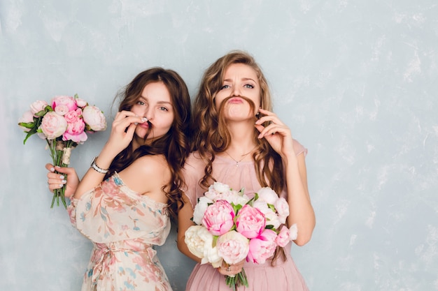 Duas lindas garotas estão em um estúdio, segurando buquês de flores e se fazendo de boba.