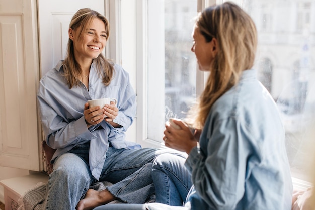 Duas lindas mulheres caucasianas, mãe e filha, estão sentadas no parapeito da janela, bebendo chá e conversando umas com as outras. Relacionamentos e conceito de tempo conjunto