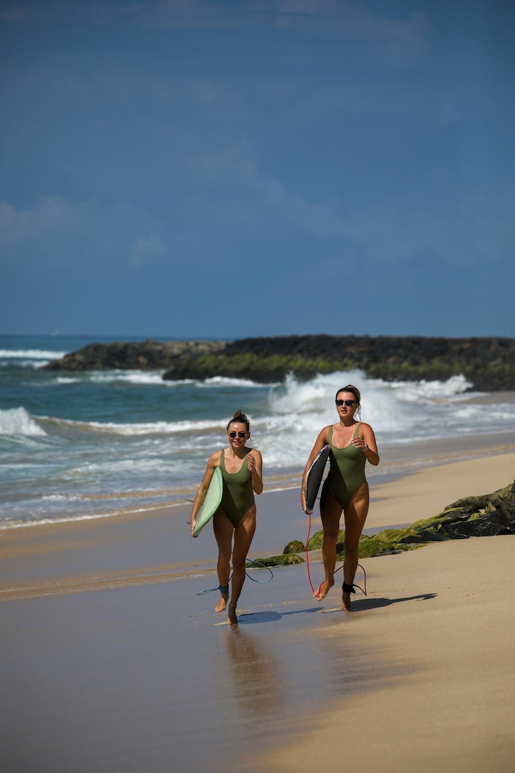 Foto grátis duas loiras bonitas em trajes de banho de uma peça correm com uma prancha de surf ao longo da praia