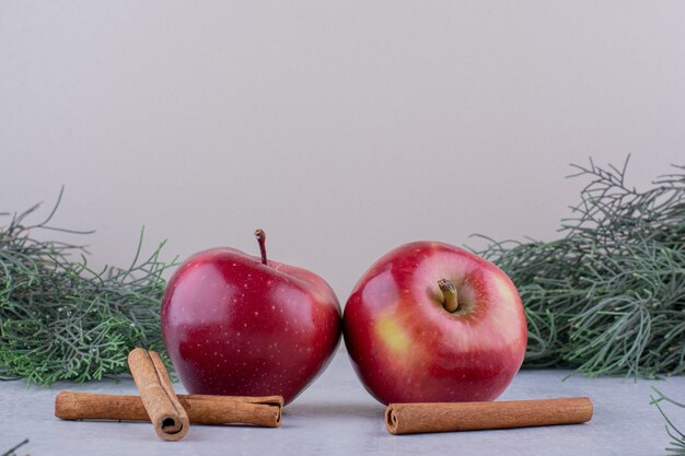 Duas maçãs e paus de canela entre ramos de pinheiro em fundo branco.