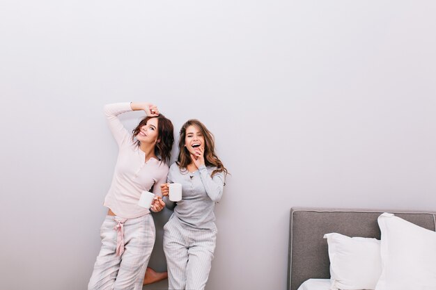 Duas meninas bonitas de pijama com xícaras, se divertindo no quarto de dormir na parede cinza.